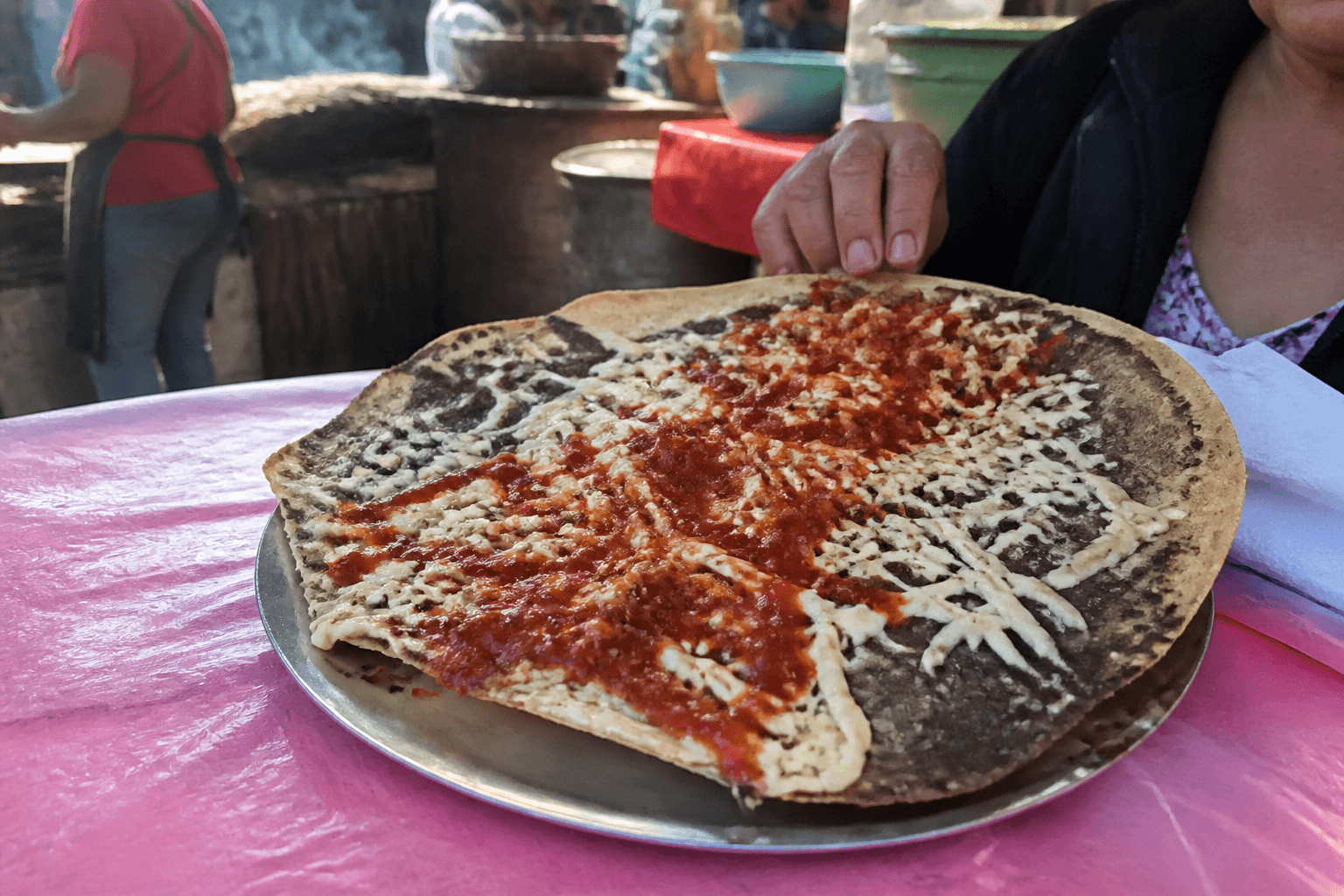 A large tlayuda with cheese and salsa on a metal plate at a Oaxaca market stall